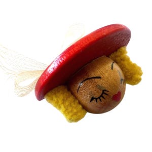 Puede incluir: Una cabeza de muñeca de madera con una cara pintada, ojos cerrados y labios rojos. La muñeca lleva un sombrero rojo y pelo de lana amarillo.