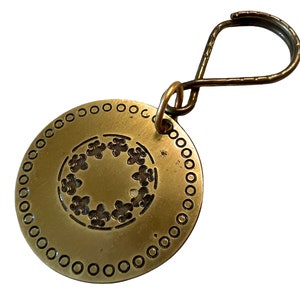 Peut inclure: Un porte-clés en laiton avec une étiquette ronde ornée d'un motif décoratif de cercle de fleurs stylisées. L'étiquette a une bordure perlée et est attachée à une simple boucle en laiton.