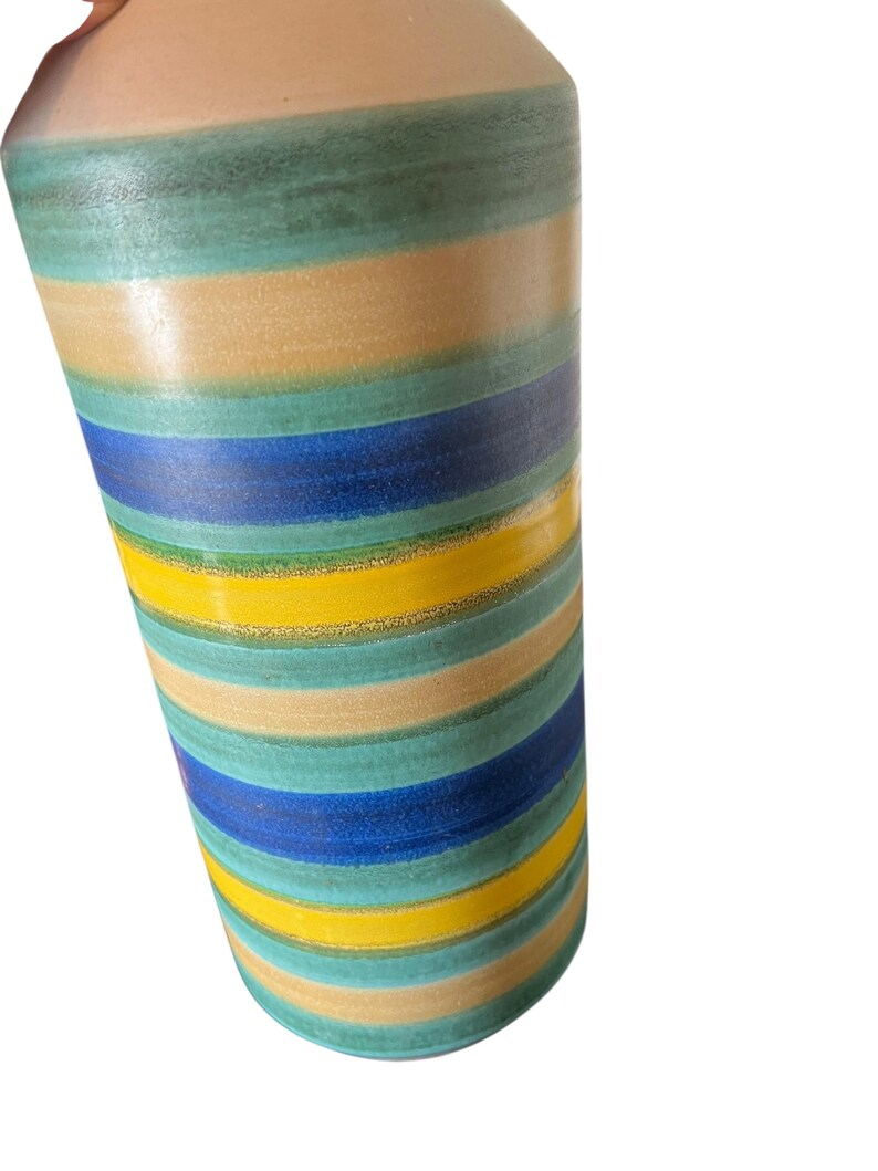 May include: A cylindrical ceramic vase with horizontal stripes in shades of blue, yellow, green, and tan. The vase has a slightly textured surface and a light beige top.