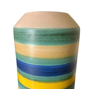 May include: A tall, cylindrical vase with horizontal stripes in shades of blue, yellow, green, and beige. The top of the vase is a solid beige color. The vase has a slightly tapered neck and a wide base.