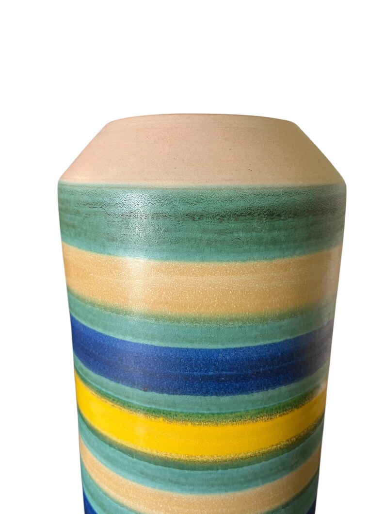 May include: A tall, cylindrical vase with horizontal stripes in shades of blue, yellow, green, and beige. The top of the vase is a solid beige color. The vase has a slightly tapered neck and a wide base.