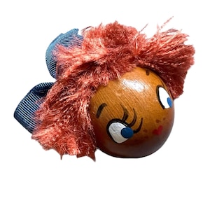 Puede incluir: Una cabeza de muñeca de madera con pelo de piel rojo, ojos azules y una cinta azul. La cabeza de la muñeca está pintada con una expresión sonriente y labios rojos. Un artículo decorativo único.