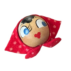 Puede incluir: Cabeza de muñeca de madera con rasgos pintados, incluyendo ojos azules, cabello negro y boca roja. La cabeza está envuelta en una bufanda de tela roja con lunares blancos. La muñeca tiene un estilo de arte popular.