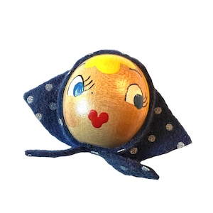 Puede incluir: Cabeza de muñeca de madera con una cara pintada, cabello rubio, ojos azules y labios rojos. La muñeca lleva un sombrero de tela azul con lunares blancos.