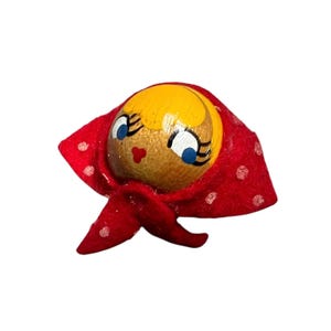 Puede incluir: Cabeza de muñeca de madera vintage con una cara pintada y un pañuelo rojo con lunares blancos. La muñeca tiene cabello rubio, ojos azules y una boca roja. La cabeza es redonda y tiene un acabado liso.