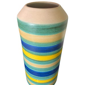 May include: A tall, cylindrical vase with a wide opening. The vase features horizontal stripes in shades of blue, yellow, green, and beige. The top rim is a solid beige color. The vase is likely made of ceramic or pottery.