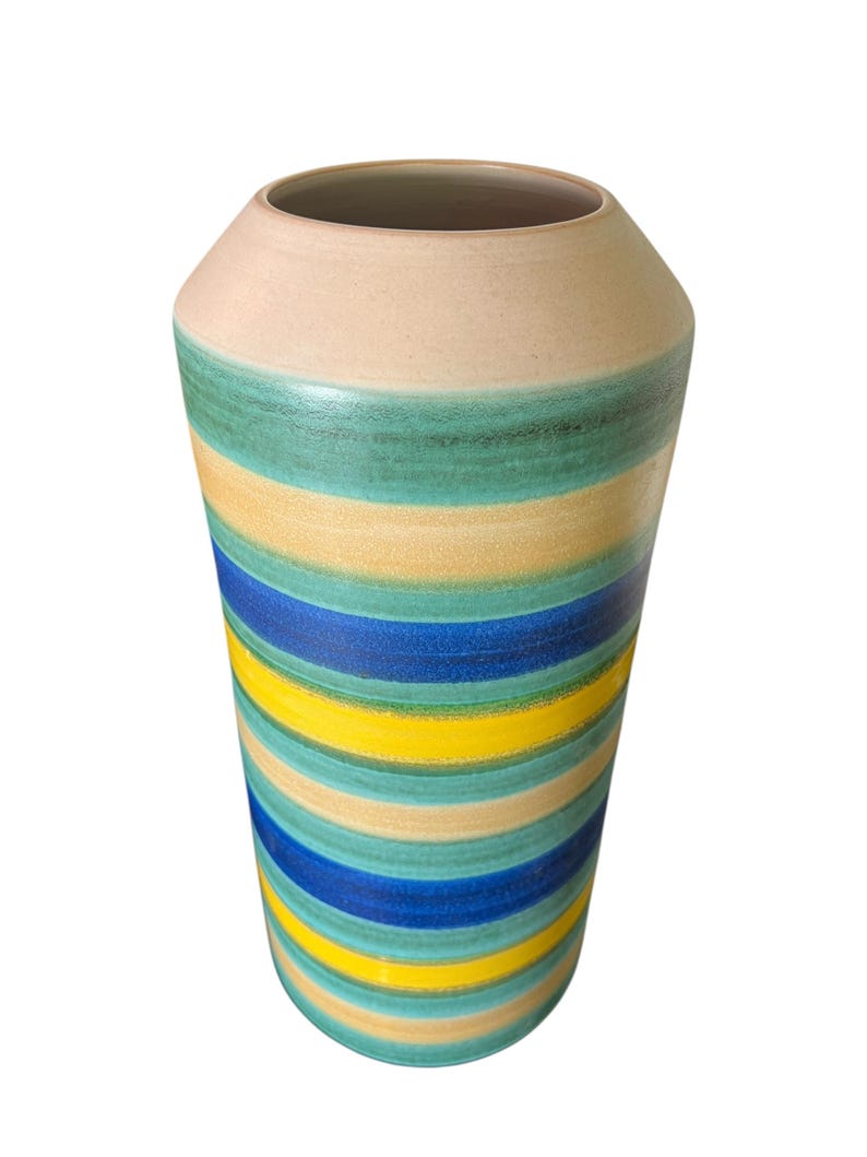 May include: A tall, cylindrical vase with a wide opening. The vase features horizontal stripes in shades of blue, yellow, green, and beige. The top rim is a solid beige color. The vase is likely made of ceramic or pottery.