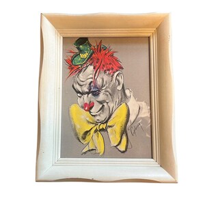 May include: Framed vintage clown portrait. The artwork features a clown with red hair, a green hat, and a large yellow bow tie. The clown's face is painted in shades of gray, with a red nose and a slight smile. The artwork is in a white frame.