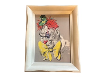 Op de afbeelding: Ingelijst vintage clownportret. Het kunstwerk toont een clown met rood haar, een groene hoed en een grote gele vlinderdas. Het gezicht van de clown is geschilderd in grijstinten, met een rode neus en een lichte glimlach. Het kunstwerk zit in een witte lijst.