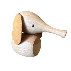 May include: A wooden elephant figurine with a light brown body, a tan ear, and a black eye. The elephant has a trunk and is made of wood. The figurine is a decorative item.
