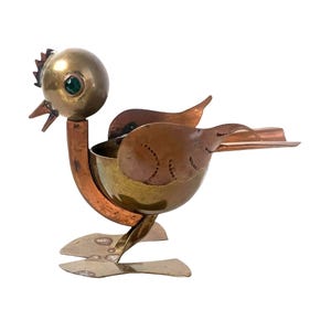 Könnte beinhalten: Ein Vintage-Aschenbecher in Vogelform aus Messing und Kupfer. Der Vogel hat einen runden Kopf mit einem grünen Edelsteinauge, einen Schnabel und einen Kamm. Der Körper hat eine Schalenform mit Flügeln und einem flachen Schwanz.