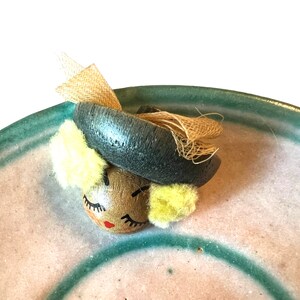 Puede incluir: Una figurita de madera de una mujer con la cara pintada, que lleva un sombrero negro con una cinta marrón y beige. La figurita está sentada en un plato de cerámica verde y rosa.