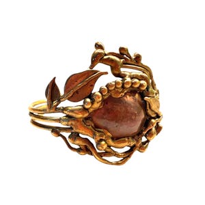 May include: A vintage-style ring with a central, reddish-brown stone, surrounded by ornate, gold-toned metalwork. The design incorporates leaf and vine motifs, creating a nature-inspired aesthetic. The ring has a multi-band design.