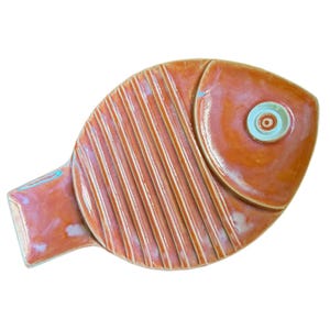 May include: A fish-shaped ceramic dish with a glossy orange glaze. The body features vertical raised ridges, while the head section is smooth and includes a circular eye detail with a light blue center and a white ring around the pupil.
