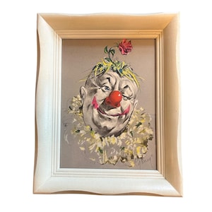 May include: A framed vintage clown portrait. The clown has a red nose, a red smile, and a ruffled collar. A red rose and green leaves adorn the top of the clown's head. The artwork is set in a white wooden frame.