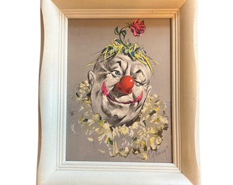 Op de afbeelding: Een ingelijst vintage clownportret. De clown heeft een rode neus, een rode glimlach en een gerimpelde kraag. Een rode roos en groene bladeren sieren de bovenkant van het hoofd van de clown. Het kunstwerk is geplaatst in een witte houten lijst.