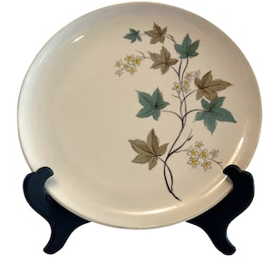 May include: A white ceramic dinner plate with a floral design featuring brown, green, and yellow leaves and flowers. The plate is on a black stand.