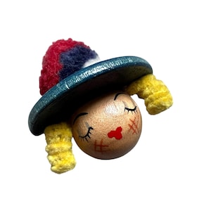 Puede incluir: Una cabeza de muñeca de madera caprichosa con ojos cerrados, labios rojos y detalles de mejillas con puntadas cruzadas. Lleva un sombrero verde azulado con un pompón rojo, blanco y azul y coletas de hilo amarillo. Un artículo decorativo único.