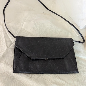 May include: A black ostrich leather clutch purse with a long strap. The purse has a flap closure and a subtle embossed pattern.