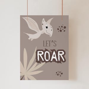 Pode incluir: Impressão emoldurada com um pterodáctilo de desenho animado e o texto "LET'S ROAR". A obra de arte usa tons de cinza e marrom, com uma samambaia estilizada e pontos espalhados. A impressão é suspensa por um fino cordão marrom.