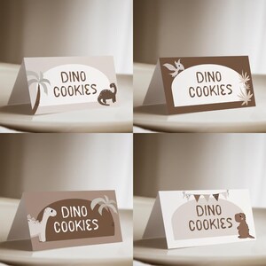 Może przedstawiać: Cztery składane karty z motywem dinozaurów. Każda karta zawiera tekst "DINO COOKIES" w białym owalu, z różnymi ilustracjami dinozaurów i elementami dekoracyjnymi w odcieniach brązu i beżu. Karty są wyświetlane na jasnej powierzchni.