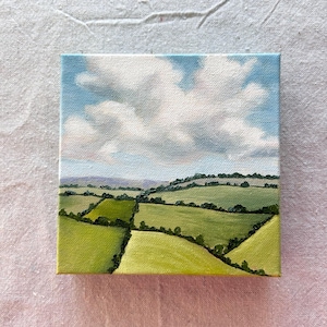 May include: A small square painting depicts a landscape with rolling green hills and fields under a blue sky with fluffy white clouds. The artwork is an oil painting on canvas, showcasing a serene countryside scene.
