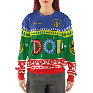 May include: A long-sleeve crewneck sweatshirt featuring a festive holiday pattern with blue, green, and red horizontal bands. The design includes white reindeer, pine trees, stars, and snowflakes. The letters DOI are printed in large, colorful font.