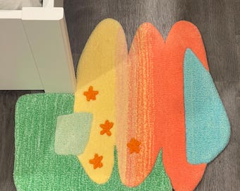 May include: A colorful, abstract rug with a green, yellow, orange, pink, and blue design. The rug has a wavy, organic shape and features small orange stars.