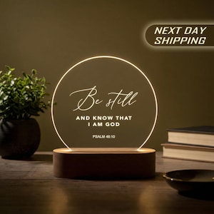 May include: A circular acrylic plaque with a warm glowing edge, mounted on a rectangular wooden base. The plaque features the white text 'Be still AND KNOW THAT I AM GOD' followed by 'PSALM 46:10' in a combination of script and sans-serif fonts.