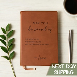 May include: A rectangular brown notebook with a leather-like texture featuring the black printed text: MAY YOU be proud of THE WORK YOU DO THE PERSON YOU ARE AND THE DIFFERENCE YOU MAKE FOR Lucia. A silver pen is positioned to the right.
