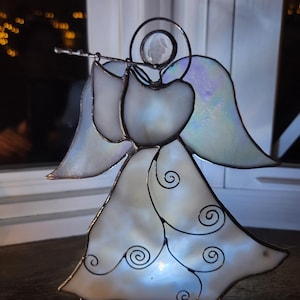 Puede incluir: Una figura de ángel de vidriera con alas iridiscentes y un halo. El ángel toca la flauta y su vestido presenta remolinos decorativos. La figura está iluminada desde dentro, creando un brillo suave. La base es de metal plateado.