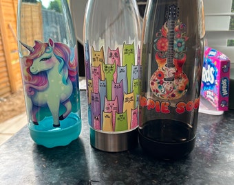 Può includere: Tre bottiglie d'acqua riutilizzabili con diversi design. La prima bottiglia è blu con un design di unicorno. La seconda bottiglia è trasparente con un design di gatti colorati. La terza bottiglia è nera con un design floreale e una chitarra.