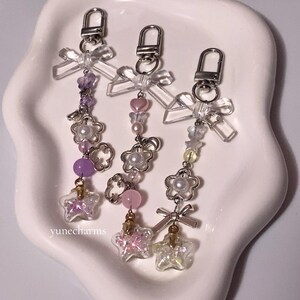 Puede incluir: Tres llaveros con anillas de plata y lazos de pl&aacute;stico transparente. Cada llavero presenta un esquema de color diferente: morado, rosa y amarillo. Los abalorios incluyen flores, cuentas y una botella con forma de estrella llena de purpurina.