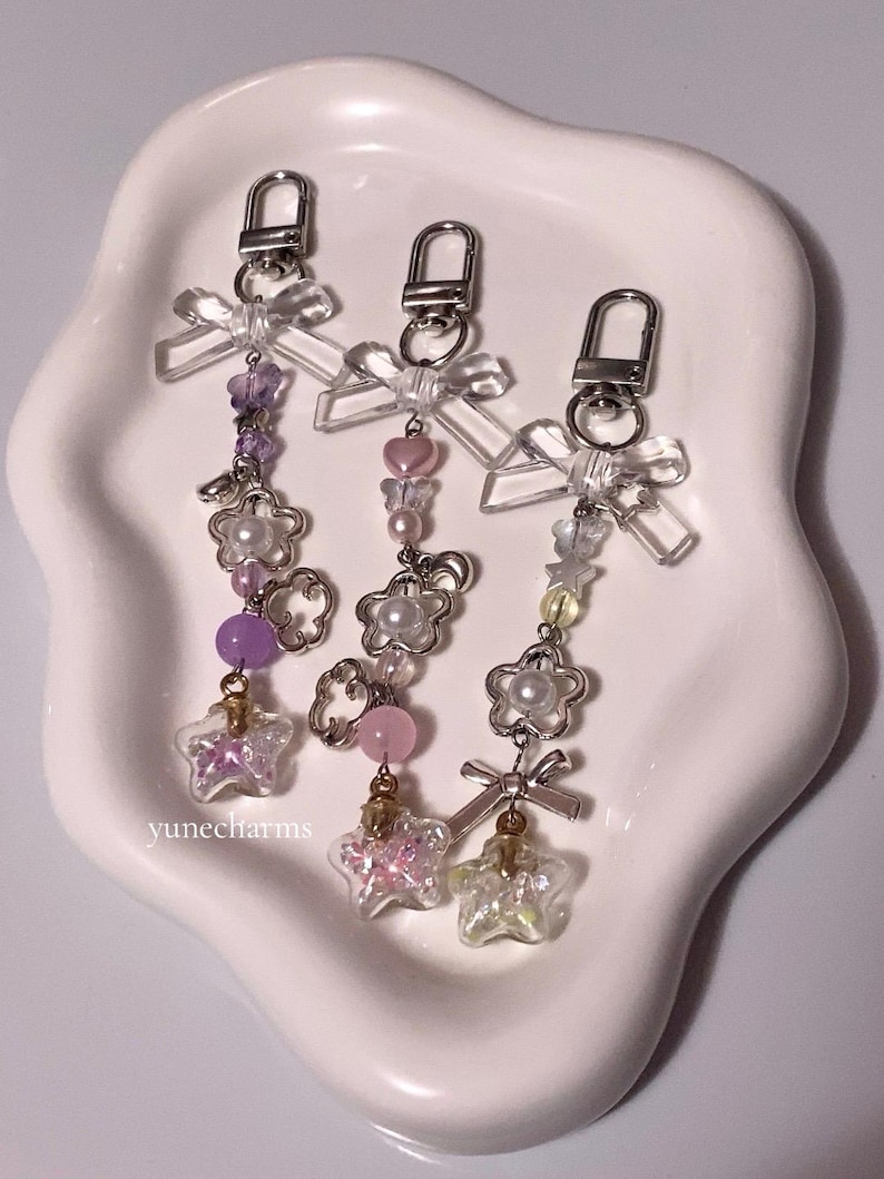 Puede incluir: Tres llaveros con anillas de plata y lazos de pl&aacute;stico transparente. Cada llavero presenta un esquema de color diferente: morado, rosa y amarillo. Los abalorios incluyen flores, cuentas y una botella con forma de estrella llena de purpurina.