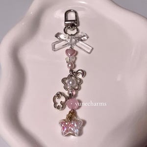 Puede incluir: Un charm rosa y blanco con un lazo transparente, un coraz&oacute;n rosa, una perla, una flor, una nube, una piedra rosa y un charm de mariposa brillante. El charm est&aacute; unido a un llavero plateado.