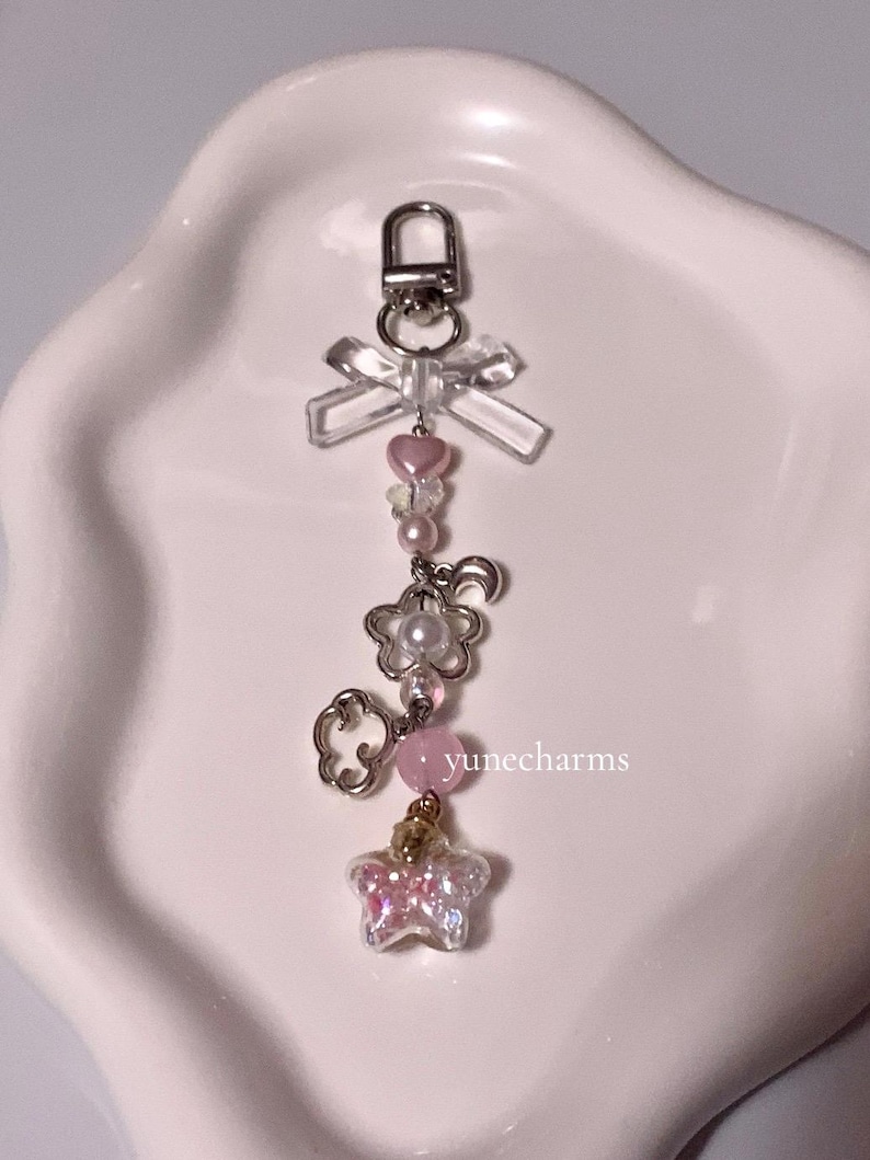 Puede incluir: Un charm rosa y blanco con un lazo transparente, un coraz&oacute;n rosa, una perla, una flor, una nube, una piedra rosa y un charm de mariposa brillante. El charm est&aacute; unido a un llavero plateado.