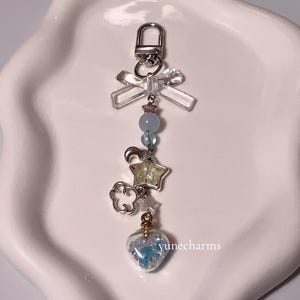 Puede incluir: Un llavero con un dije de plata, un lazo transparente, cuentas azules, una estrella plateada, una nube plateada y un dije en forma de coraz&oacute;n lleno de brillantina azul.