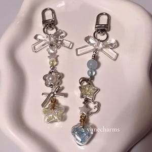 Puede incluir: Dos llaveros plateados con lazos de cristal transparente, cuentas y dijes. Un llavero tiene un dije de coraz&oacute;n azul, el otro tiene un dije de estrella amarilla. Los dijes est&aacute;n sobre un fondo blanco. El texto "yunecharms" es visible en la esquina inferior derecha.