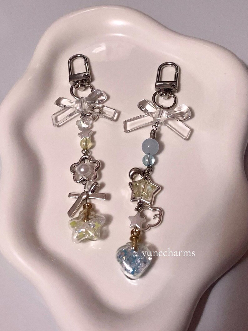 Puede incluir: Dos llaveros plateados con lazos de cristal transparente, cuentas y dijes. Un llavero tiene un dije de coraz&oacute;n azul, el otro tiene un dije de estrella amarilla. Los dijes est&aacute;n sobre un fondo blanco. El texto "yunecharms" es visible en la esquina inferior derecha.