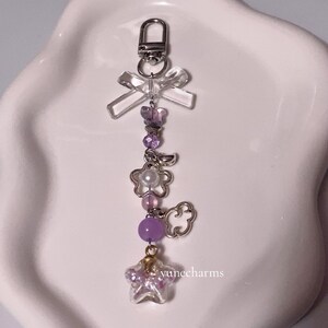 Puede incluir: Un llavero con charms morado y blanco, que incluye un charm de estrella transparente, un charm de nube, un charm de flor y un charm de lazo transparente. El llavero est&aacute; unido a un cierre de metal plateado.