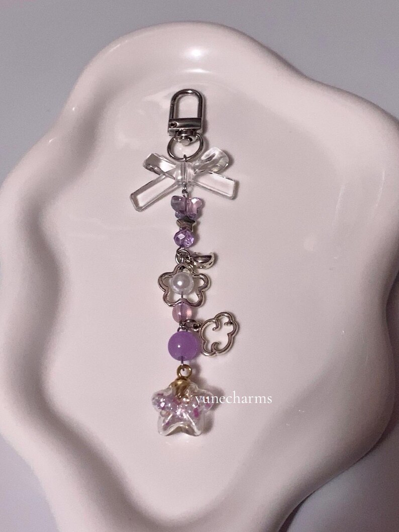 Puede incluir: Un llavero con charms morado y blanco, que incluye un charm de estrella transparente, un charm de nube, un charm de flor y un charm de lazo transparente. El llavero est&aacute; unido a un cierre de metal plateado.