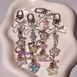 Puede incluir: Cuatro llaveros con dijes, cierres de metal plateado y botellas de vidrio transparente llenas de brillantina y peque&ntilde;os dijes. Los dijes incluyen estrellas, corazones, lunas y flores. Los llaveros est&aacute;n sobre una superficie blanca.