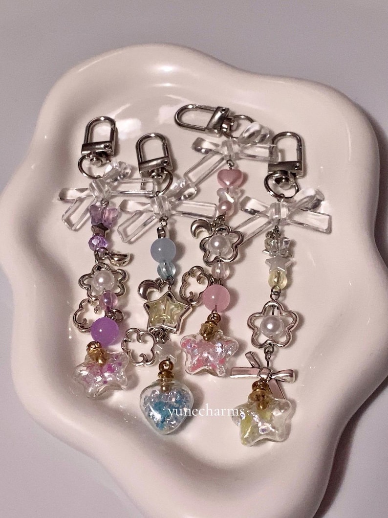 Puede incluir: Cuatro llaveros con dijes, cierres de metal plateado y botellas de vidrio transparente llenas de brillantina y peque&ntilde;os dijes. Los dijes incluyen estrellas, corazones, lunas y flores. Los llaveros est&aacute;n sobre una superficie blanca.