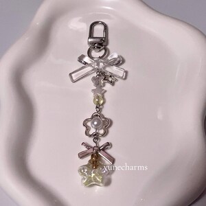 Puede incluir: Un llavero de plata con un lazo transparente, un dije de flor de perla y un dije de estrella amarilla. El llavero tiene un cierre de plata.