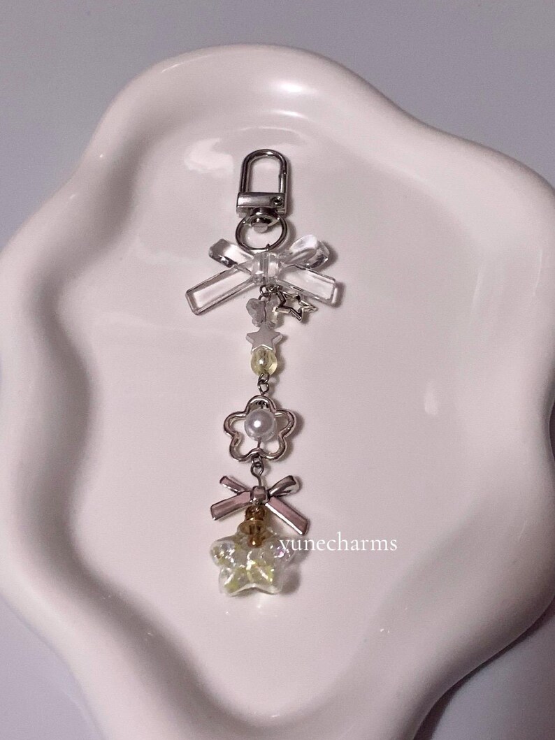 Puede incluir: Un llavero de plata con un lazo transparente, un dije de flor de perla y un dije de estrella amarilla. El llavero tiene un cierre de plata.