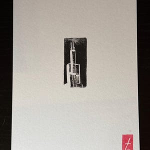 Peut inclure: Une carte rectangulaire blanche présentant une impression en bloc d'encre noire centrée représentant la silhouette d'un gratte-ciel. Dans le coin inférieur gauche, le texte 05/08 est écrit à l'encre noire. Un petit carré rouge avec un t blanc est en bas.