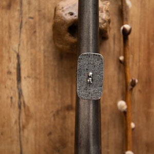 Puede incluir: Un anillo de plata con una banda rectangular texturizada y dos pequeñas cuentas de plata. El anillo se muestra sobre una superficie de madera.