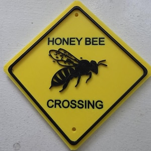 Può includere: Un cartello giallo a forma di rombo con bordo nero e due fori di montaggio. Il cartello presenta al centro la sagoma nera di un'ape, con il testo nero HONEY BEE sopra e CROSSING sotto la grafica sulla superficie gialla.