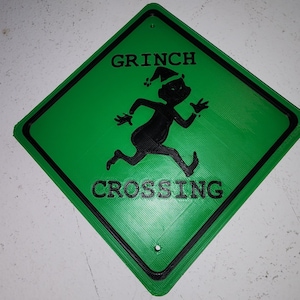 May include: A diamond-shaped green sign with a black border and two mounting holes. The sign features a black silhouette of a running character wearing a hat, with the black text GRINCH above and the black text CROSSING below the figure.