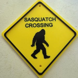 May include: A diamond-shaped yellow sign with a black border and a black silhouette of a Sasquatch walking. The text SASQUATCH CROSSING is printed in black capital letters above the figure. There are two small circular holes at the top and bottom.